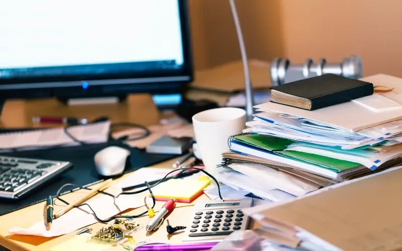 A disorganized desk with a computer on it and a stack of papers with Bible atop it.